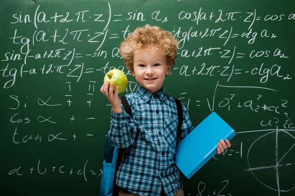 Cheerful kid holding apple and book near chalkboard with mathematical formulas — Stock Photo