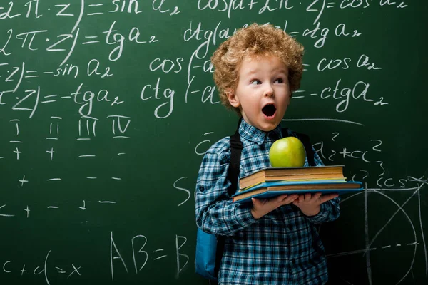 Niño sorprendido sosteniendo libros y manzana cerca de pizarra con fórmulas matemáticas - foto de stock