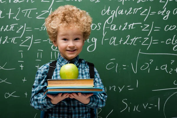 Niño rizado sosteniendo libros y manzana cerca de pizarra con fórmulas matemáticas - foto de stock