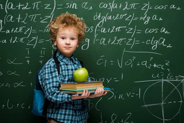 Bambino riccio che tiene libri e mela fresca vicino alla lavagna con formule matematiche — Foto stock