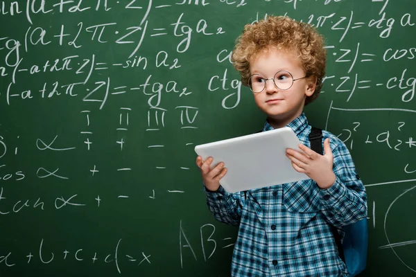 Smart kid in glasses holding digital tablet near chalkboard with mathematical formulas — Stock Photo
