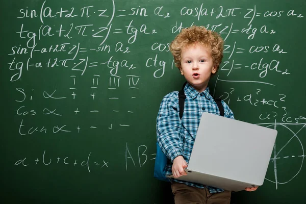 Garoto surpreso segurando laptop perto de quadro-negro com fórmulas matemáticas — Fotografia de Stock