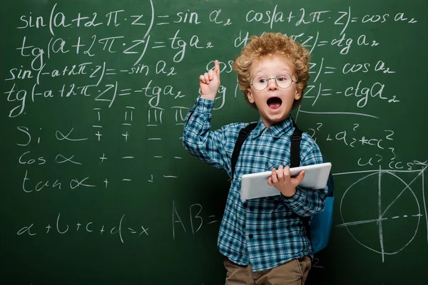 Schlaues Kind mit Brille hat Idee, während es digitales Tablet in der Nähe der Tafel hält — Stockfoto