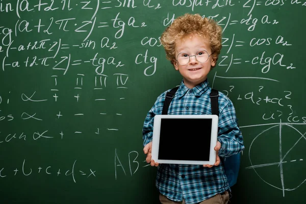 Niño feliz e inteligente en gafas que sostienen la tableta digital con pantalla en blanco cerca de pizarra con fórmulas matemáticas - foto de stock