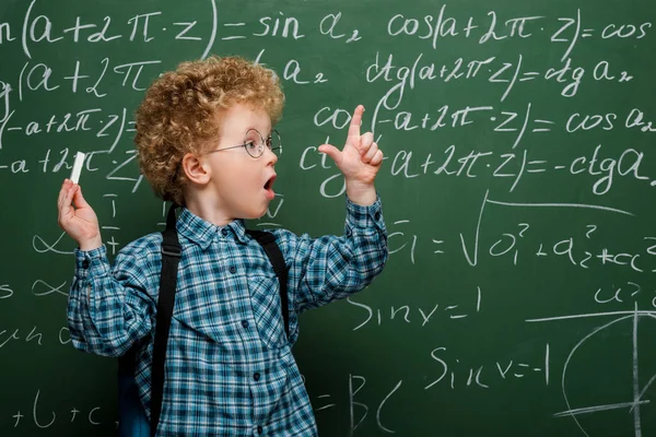 Enfant surpris dans des lunettes geste tout en tenant la craie près du tableau avec des formules mathématiques — Photo de stock