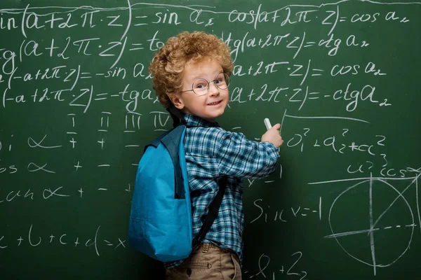 Happy kid in glasses writing mathematical formulas on chalkboard — Stock Photo