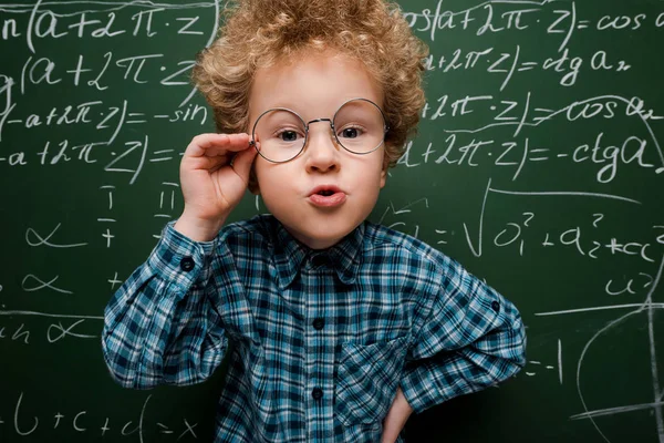 Niño inteligente tocando gafas y de pie con la mano en la cadera cerca de pizarra - foto de stock