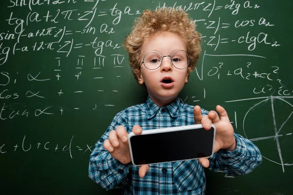 Smart kid in glasses holding smartphone with blank screen near chalkboard with mathematical formulas — Stock Photo