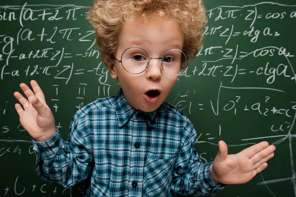 Surprised kid in glasses looking at camera near chalkboard with mathematical formulas — Stock Photo