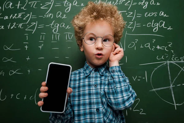 Smart child holding smartphone with blank screen and touching glasses near chalkboard with mathematical formulas — Stock Photo