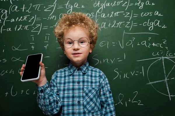 Inteligente niño sosteniendo teléfono inteligente con pantalla en blanco cerca de pizarra con fórmulas matemáticas - foto de stock