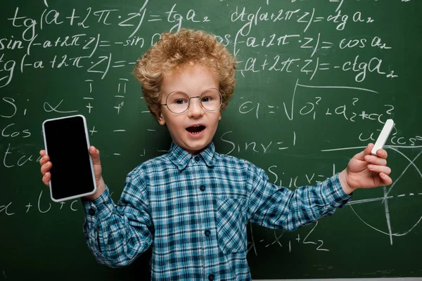 Mignon et intelligent enfant tenant smartphone avec écran vierge et tenant craie près de tableau noir avec des formules mathématiques — Photo de stock