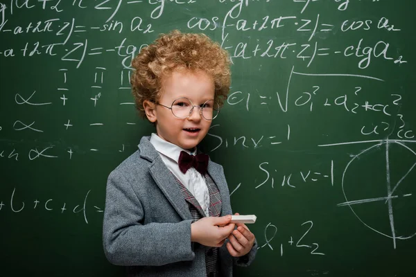 Niño lindo e inteligente en traje con corbata de lazo sosteniendo tiza cerca de pizarra con fórmulas matemáticas - foto de stock