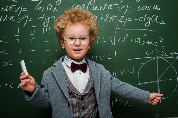 Happy kid in suit with bow tie holding chalk near chalkboard with mathematical formulas — Stock Photo