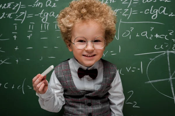 Niño inteligente con traje y pajarita sosteniendo tiza cerca de pizarra con fórmulas matemáticas - foto de stock