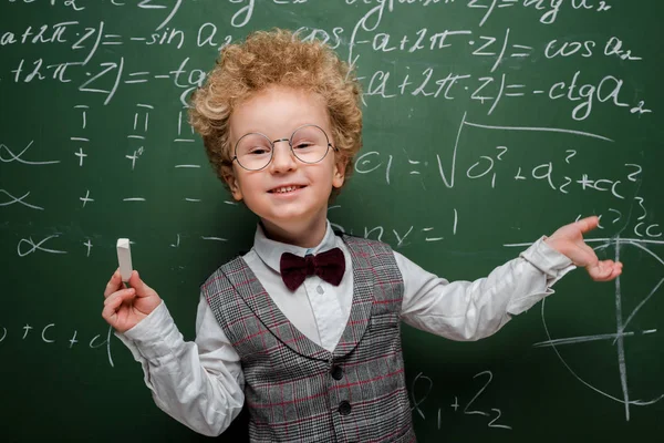 Bambino sorridente in tuta e papillon tenendo il gesso e puntando con mano alla lavagna con formule matematiche — Foto stock