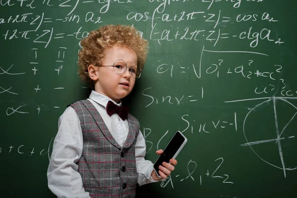 Niño disgustado con traje y pajarita sosteniendo teléfono inteligente con pantalla en blanco cerca de pizarra con fórmulas matemáticas - foto de stock