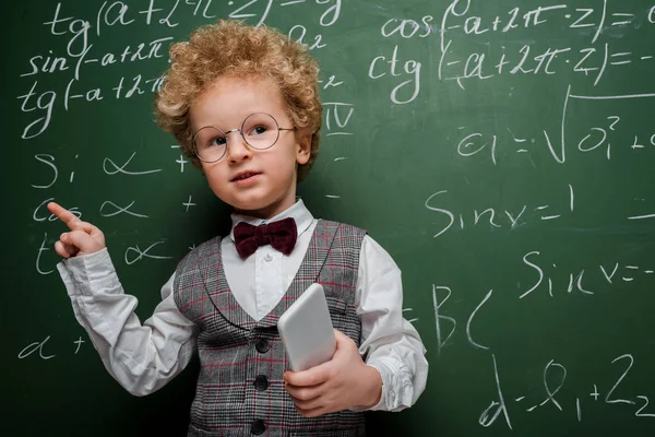 Enfant intelligent en costume et noeud papillon tenant smartphone et pointant du doigt près du tableau avec des formules mathématiques — Photo de stock