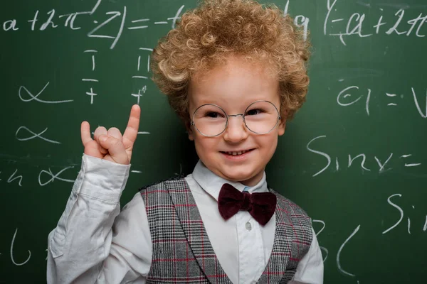 Niño feliz e inteligente en traje y pajarita mostrando letrero de roca cerca de pizarra con fórmulas matemáticas - foto de stock