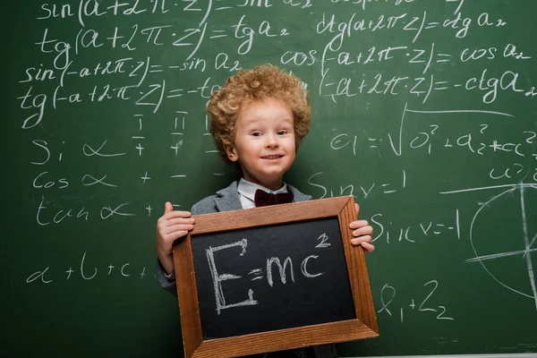 Criança feliz em terno e gravata arco segurando pequeno quadro negro com fórmula perto de quadro-negro — Fotografia de Stock