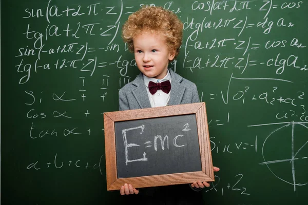 Smart child in suit and bow tie holding small blackboard with formula near chalkboard — Stock Photo