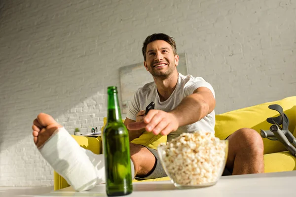 Foco seletivo de homem sorridente com perna quebrada assistindo tv perto de garrafa de cerveja e pipoca na sala de estar — Fotografia de Stock