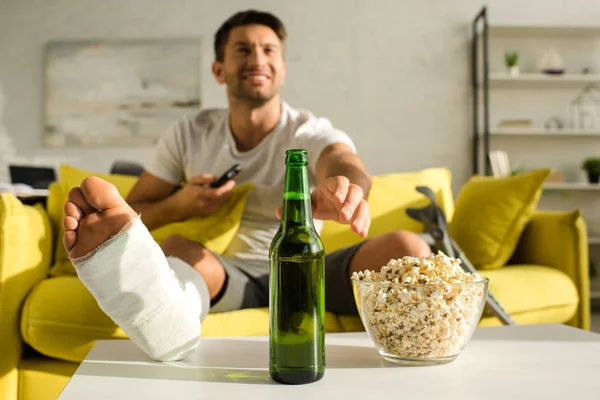 Selective focus of smiling man with broken leg taking beer near popcorn while watching tv on couch — Stock Photo