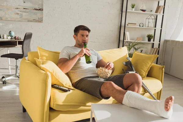 Schöner Mann mit gebrochenem Bein hält Popcorn und Bier in der Hand, während er im Wohnzimmer fernsieht — Stockfoto