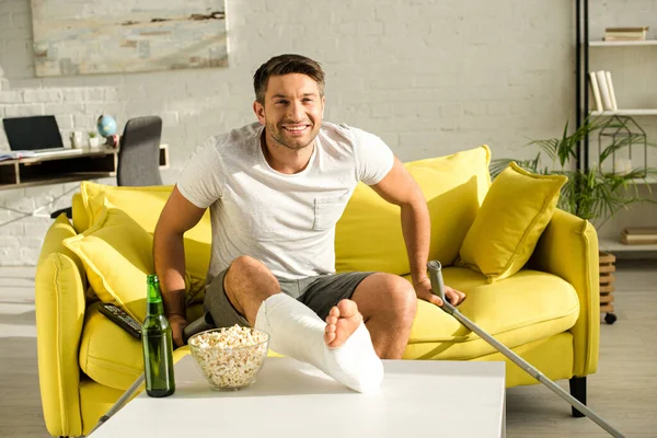 Homme souriant avec jambe cassée regardant la caméra près de la bouteille de bière et du maïs soufflé sur la table basse dans le salon — Photo de stock