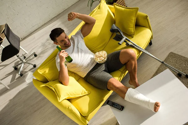 Vue grand angle de l'homme gai avec jambe cassée regarder la télévision avec du maïs soufflé et de la bière sur le canapé — Photo de stock