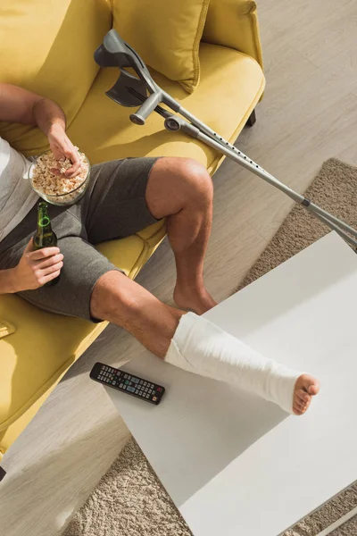 Vista superior do homem com a perna quebrada na mesa de café beber cerveja e comer pipocas no sofá em casa — Fotografia de Stock