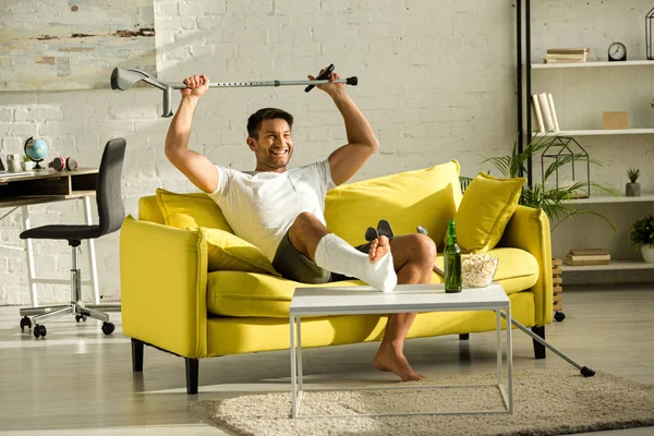 Cheerful man with broken leg raising crutch near beer bottle and popcorn on couch — Stock Photo