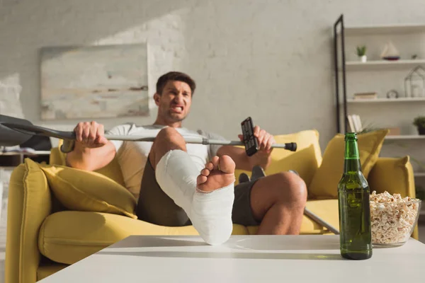 Concentration sélective de l'homme en colère avec jambe cassée et télécommande assis sur le canapé près de la bière et du maïs soufflé sur la table basse — Photo de stock