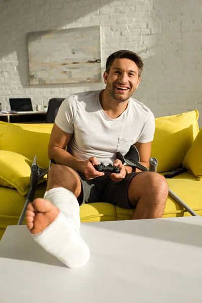 QUIIV, UCRÂNIA - JANEIRO 21, 2020: Foco seletivo de homem sorridente com perna quebrada segurando joystick e olhando para a câmera no sofá — Fotografia de Stock