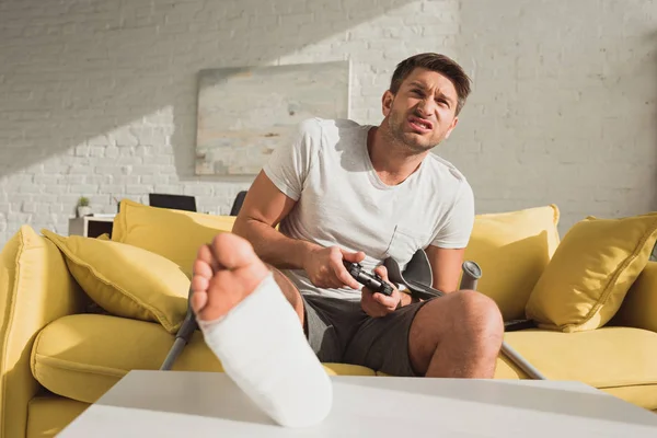 KYIV, UKRAINE - JANUARY 21, 2020: Selective focus of angry man with broken leg playing video game on sofa at home — Stock Photo