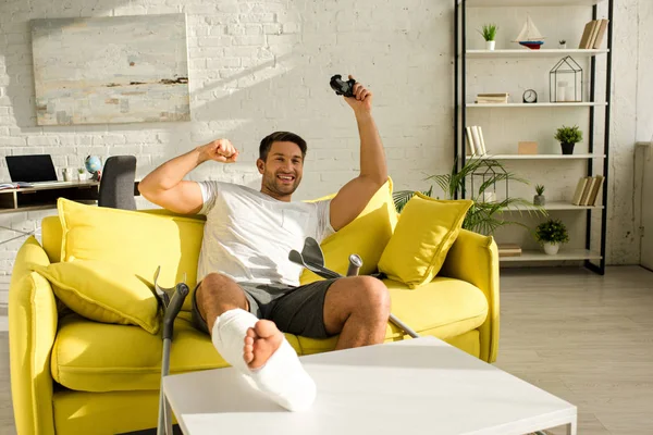 KYIV, UKRAINE - 21 JANVIER 2020 : Homme joyeux avec la jambe cassée souriant tout en tenant joystick sur le canapé à la maison — Photo de stock