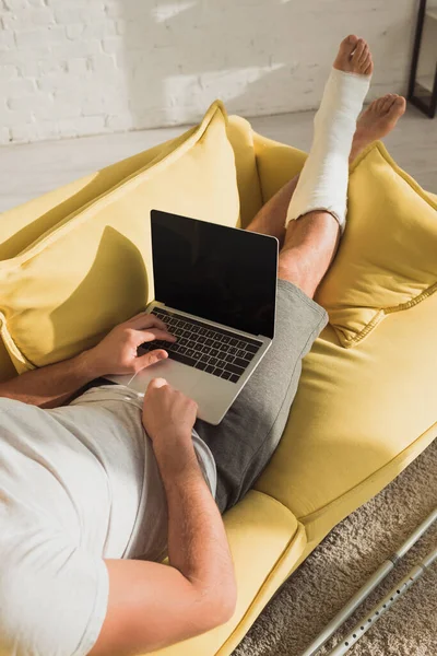 Ausgeschnittene Ansicht eines Mannes mit gebrochenem Bein tippt auf Laptop auf Sofa — Stockfoto