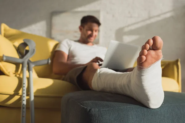 Selective focus of freelancer with broken leg on ottoman using laptop on couch — Stock Photo