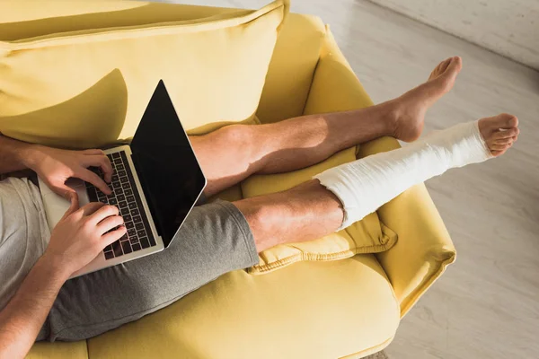 Vista recortada del freelancer con pierna en venda de yeso usando portátil en sofá en casa - foto de stock