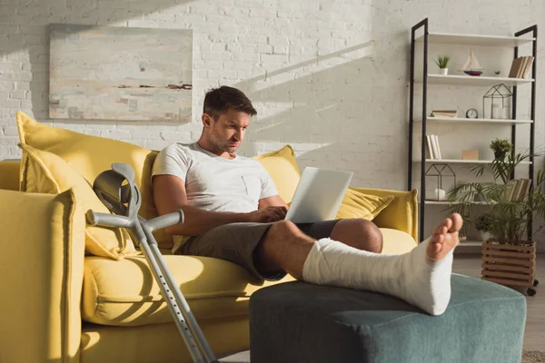 Schöner Mann mit gebrochenem Bein mit Laptop in der Nähe von Krücken auf Sofa — Stockfoto