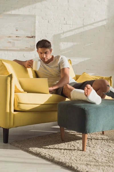 Beau homme avec jambe cassée en utilisant un ordinateur portable et tenant la tasse sur le canapé — Photo de stock