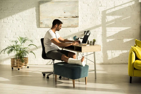 Vista lateral del freelancer con la pierna rota trabajando en el portátil en la sala de estar - foto de stock