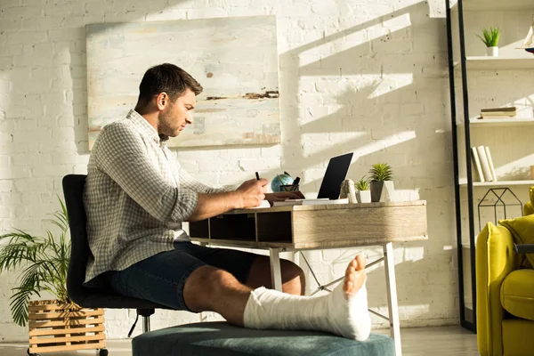 Vista laterale del bel freelance con gamba rotta che scrive sul taccuino e che usa il computer portatile a tavola — Foto stock