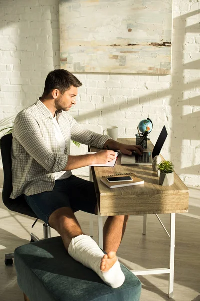 Vue latérale du pigiste avec jambe cassée à l'aide d'un ordinateur portable près des ordinateurs portables et smartphone sur la table — Photo de stock