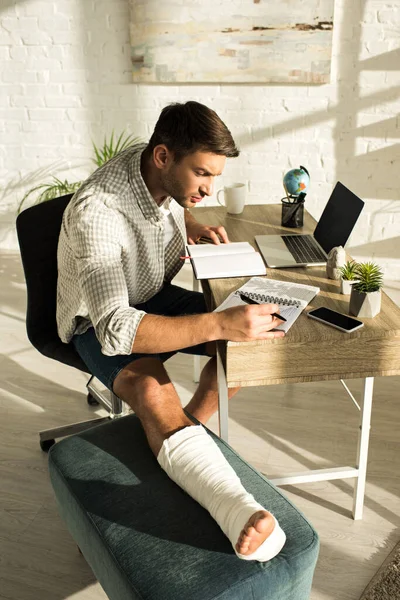 Vista lateral del apuesto freelancer con la pierna rota trabajando con cuadernos y portátil en la mesa - foto de stock