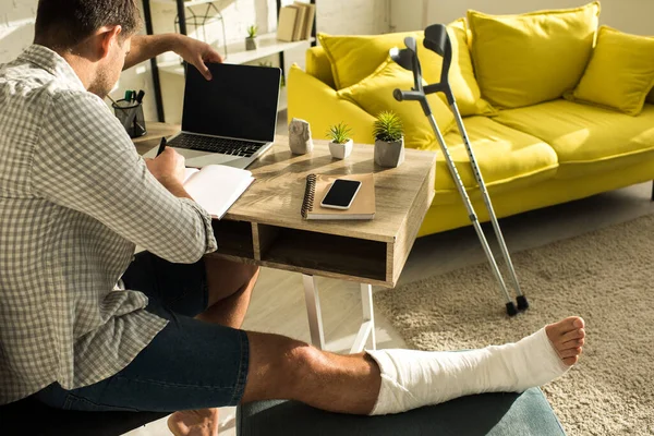 Back view of freelancer with broken leg working with laptop and notebook at table — Stock Photo