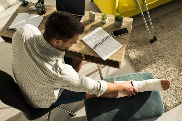 Overhead view of freelancer touching broken leg while sitting at table with laptop and notebook — Stock Photo