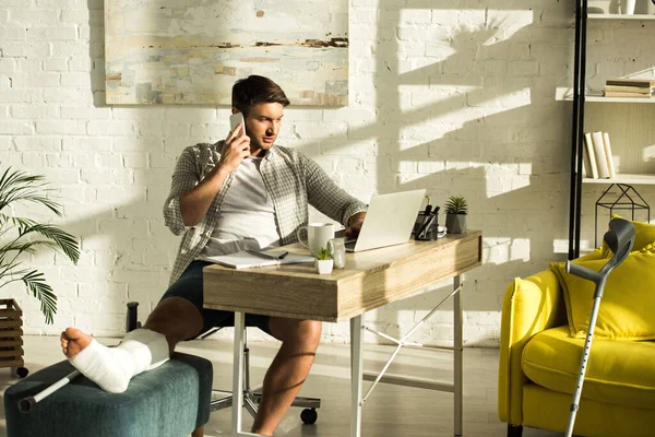 Freelancer bonito com perna quebrada usando laptop e falando no smartphone à mesa na sala de estar — Fotografia de Stock