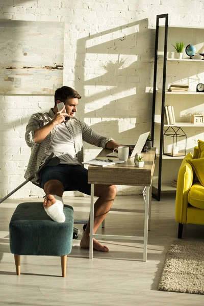 Freelancer con la pierna rota usando portátil y hablando en smartphone en la mesa en la sala de estar - foto de stock