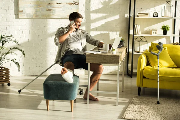 Beau pigiste avec jambe cassée sur ottoman parlant sur smartphone et utilisant un ordinateur portable à la table — Photo de stock